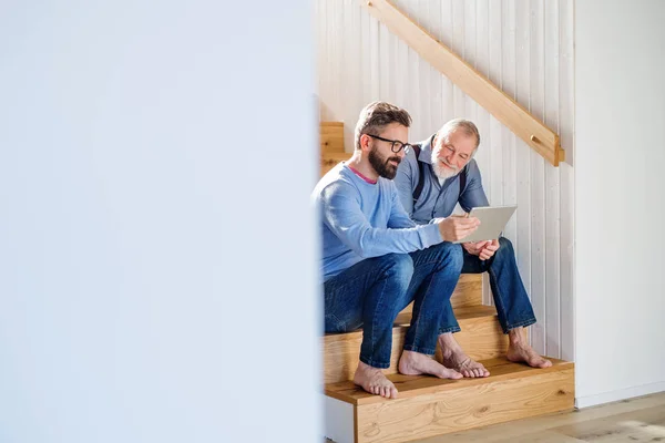 Ein erwachsener Sohn und ein älterer Vater mit Tablet sitzen zu Hause auf der Treppe. — Stockfoto