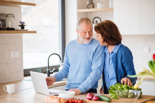 Ett porträtt av Senior par med laptop inomhus hemma, Matlagning. — Stockfoto