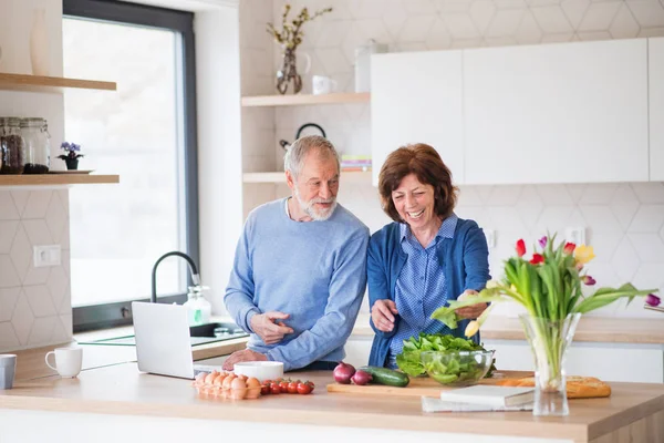 Um retrato de casal sênior com laptop dentro de casa, cozinhar . — Fotografia de Stock