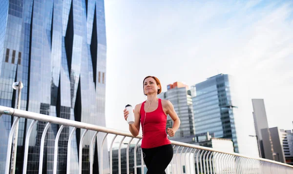 市内でイヤホンと水筒ジョギングをする若い女性ランナー. — ストック写真