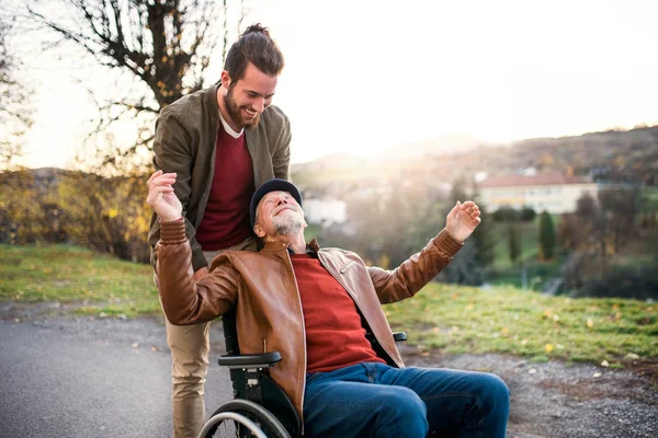 Młody mężczyzna i jego starszy ojciec na wózku inwalidzkim na spacerze w mieście. — Zdjęcie stockowe