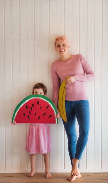 Uma jovem mulher com filha pequena dentro de casa, segurando frutas de brinquedo . — Fotografia de Stock
