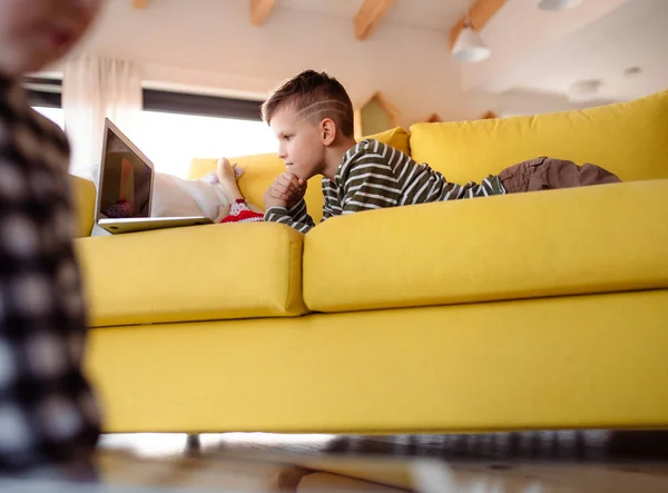 Mały chłopiec leżący w pomieszczeniu na kanapie w domu, za pomocą laptopa. — Zdjęcie stockowe