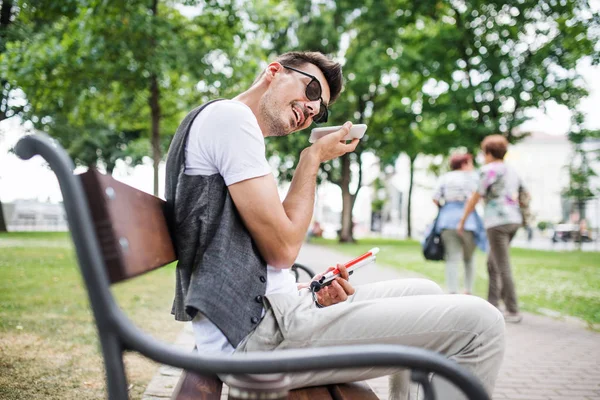 Ung blind man med smartphone sitter på bänken i parken i staden. — Stockfoto