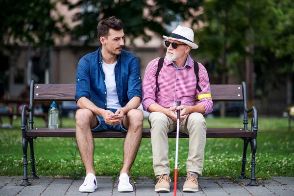 Jovem e cego sênior com cana branca sentado no banco no parque na cidade . — Fotografia de Stock