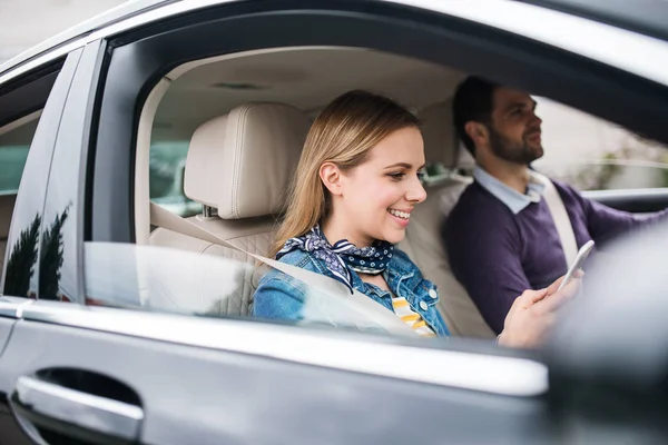 Feliz joven pareja sentada en el coche, utilizando el teléfono inteligente . — Foto de Stock