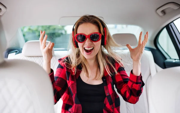 Mladá žena s brýlemi a sluchátky v autě, se zábavou. — Stock fotografie