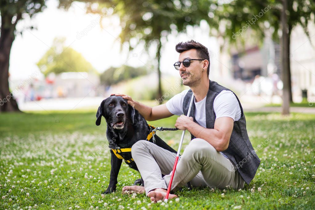 Young blind man with white cane and guide dog sitting in park in city.