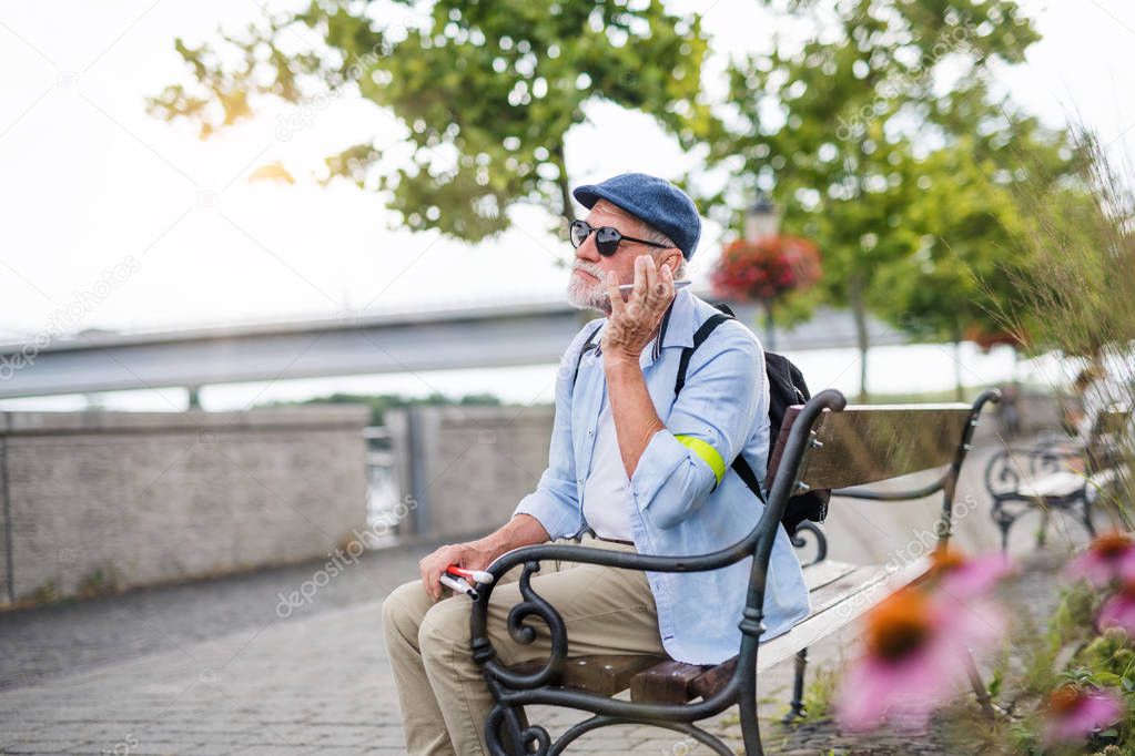 Senior blind man with smartphone sitting on bench in park in city.