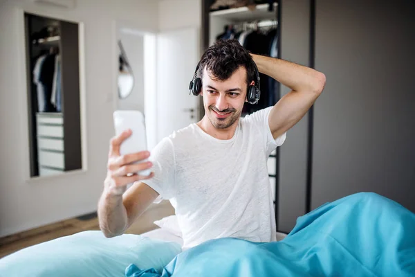 Молодой человек с наушниками и смартфоном в постели дома, слушает музыку . — стоковое фото