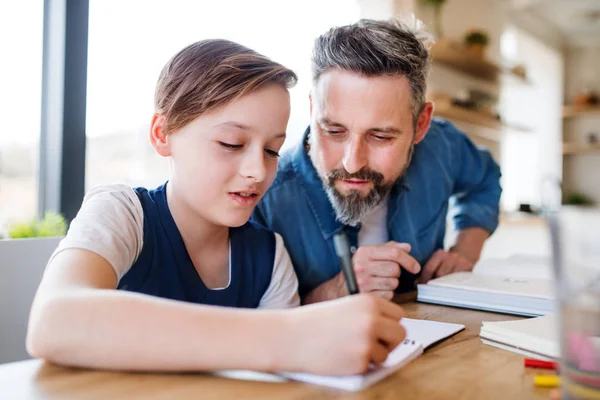 Volwassen vader met kleine zoon zittend aan tafel binnenshuis, het doen van huiswerk. — Stockfoto