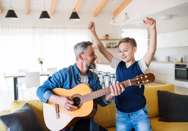Dojrzały ojciec z małym synem siedzi na kanapie w pomieszczeniu, grając na gitarze. — Zdjęcie stockowe