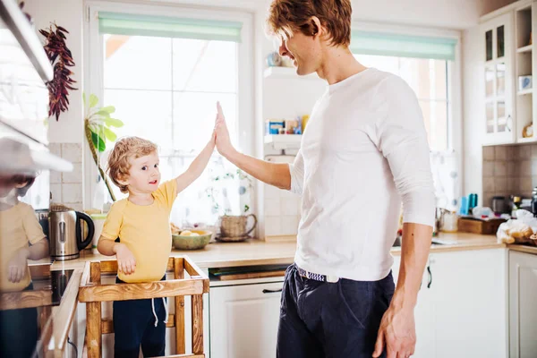 Um jovem pai com um filho menor em uma cozinha em casa, dando mais cinco . — Fotografia de Stock