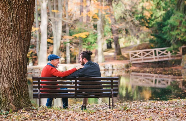 Visão traseira do pai sênior e seu filho sentado no banco por lago na natureza, falando . — Fotografia de Stock