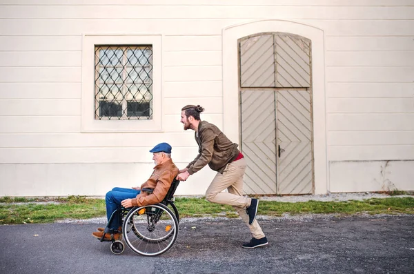 Young man and his senior father in wheelchair on a walk in town, having fun. — Stock Photo, Image