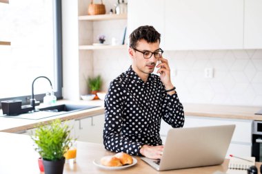 Dizüstü bilgisayar ve akıllı telefon mutfakta oturan genç adam, bir ev ofis konsepti.
