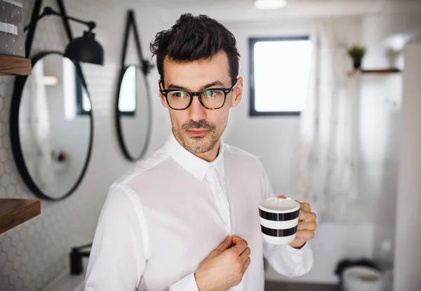 Young man with white shirt in the bathroom in the morning, holding a cup of coffee.