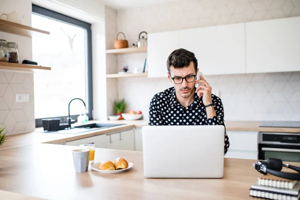 キッチンに座ってラップトップとスマートフォンを持つ若い男、ホームオフィスのコンセプト. — ストック写真