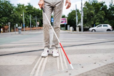 Midsection of young blind man with white cane walking across the street in city. clipart