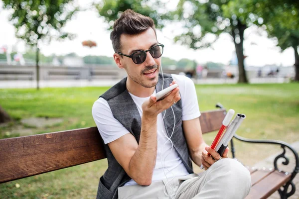 Ung blind man med smartphone sitter på bänken i Park i staden, ringer. — Stockfoto