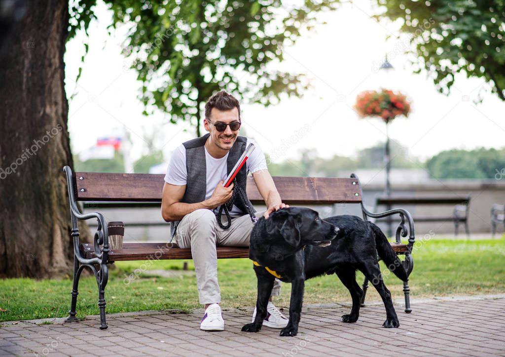 Young blind man with white cane and guide dog sitting in park in city.