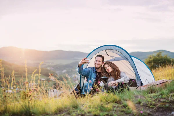 Jovens viajantes casal turístico com tenda abrigo sentado na natureza, tomando selfie . — Fotografia de Stock