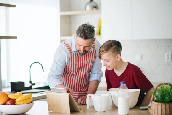 Mature father and small son with tablet indoors in kitchen, making pancakes. — Stock Photo, Image