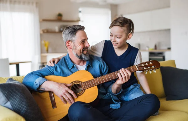 Dojrzały ojciec z małym synem siedzi na kanapie w pomieszczeniu, grając na gitarze. — Zdjęcie stockowe