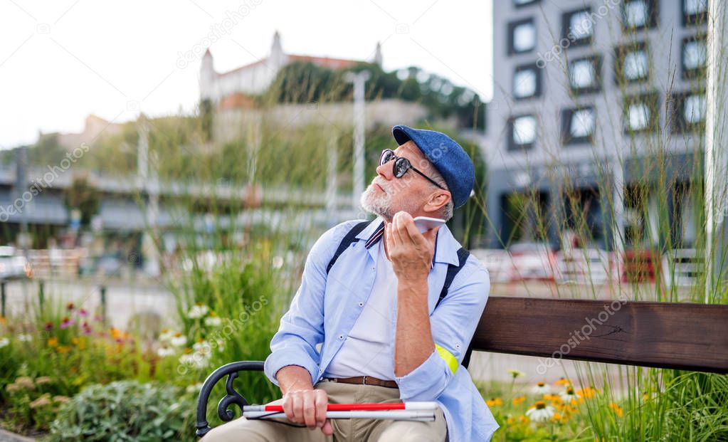 Senior blind man with smartphone sitting on bench in park in city.