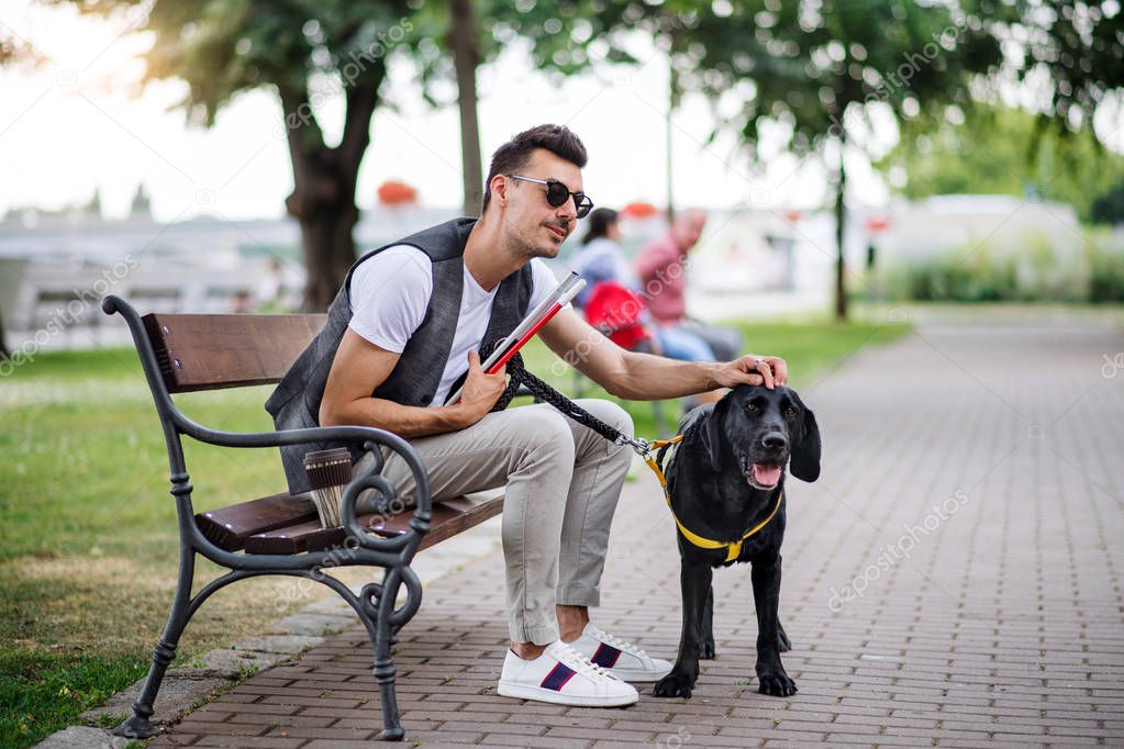 Young blind man with white cane and guide dog sitting in park in city.