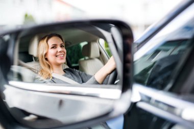 Genç kadın sürücünün arabada oturup araba sürüşünün ayna yansıması..
