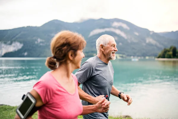 Senior pensioner couple with smartphone running by lake in nature.