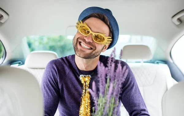 Homme joyeux avec des accessoires de fête assis dans la voiture, s'amuser . — Photo