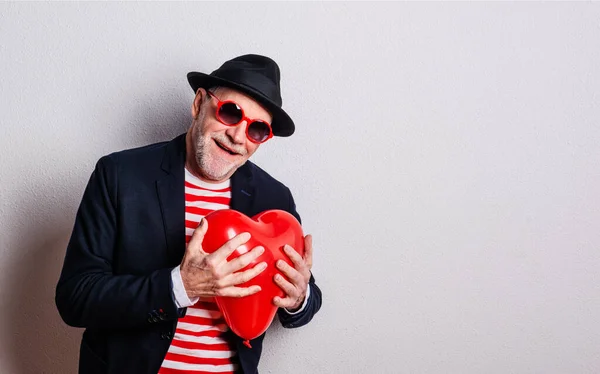 Retrato de um homem idoso apaixonado em um estúdio, segurando um balão de coração vermelho . — Fotografia de Stock