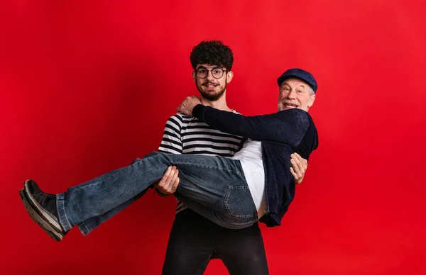Ung man som håller äldre far i en studio på röd bakgrund. — Stockfoto