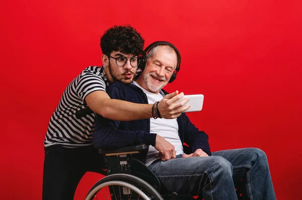 Giovane e padre anziano in sedia a rotelle in uno studio su sfondo rosso, scattando selfie . — Foto Stock