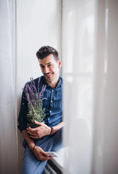 Um retrato de um jovem sentado no peitoril da janela dentro de casa, segurando uma planta . — Fotografia de Stock