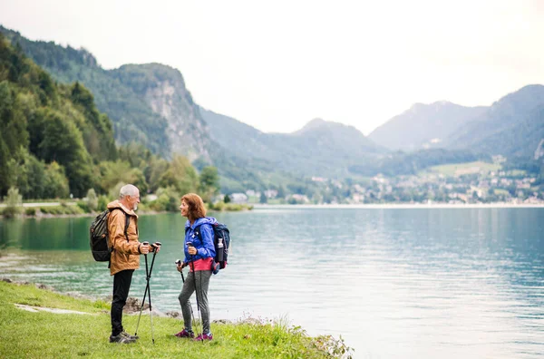 Um casal de pensionistas seniores caminhantes em pé junto ao lago na natureza, conversando . — Fotografia de Stock