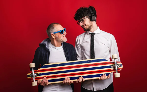 Młody mężczyzna i starszy ojciec z longboard w studio na czerwonym tle. — Zdjęcie stockowe