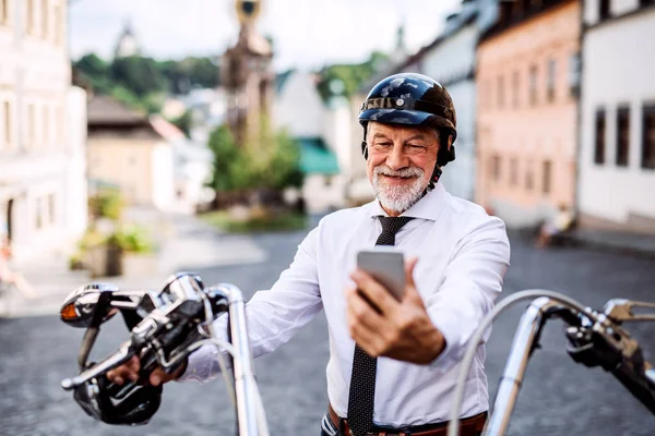 Un homme d'affaires senior avec une moto en ville, en utilisant un smartphone . — Photo