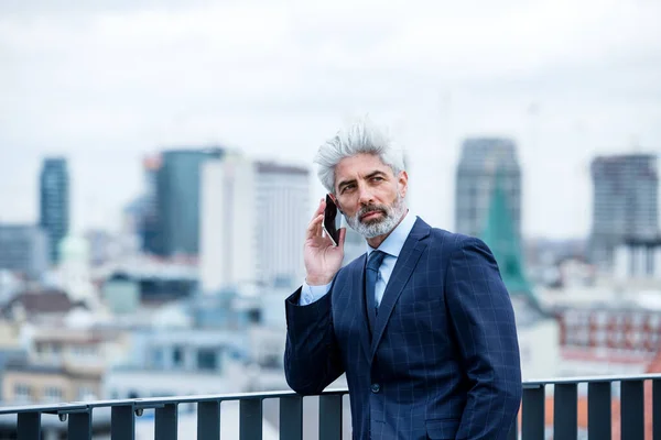Un uomo d'affari maturo con smartphone in piedi su una terrazza, al lavoro . — Foto Stock
