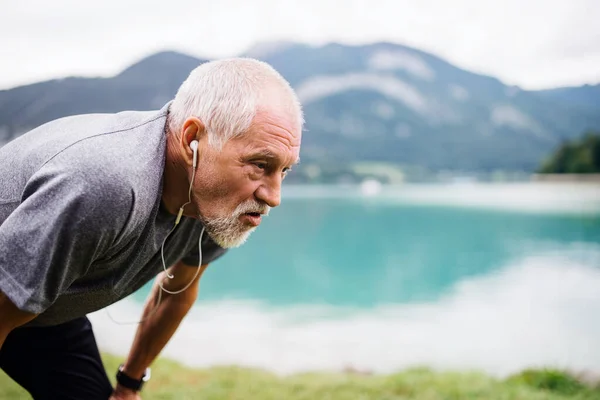 Um homem sênior com fones de ouvido em pé junto ao lago na natureza, fazendo exercício . — Fotografia de Stock