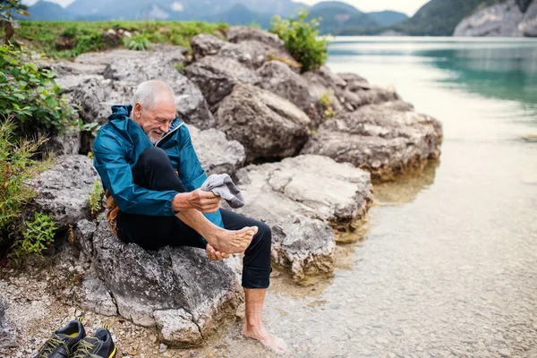 Starszy człowiek spacerowicz siedzi nad jeziorem w naturze, zdejmuje buty. — Zdjęcie stockowe