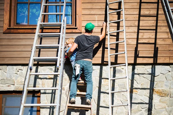 Un père et un tout-petit garçon à l'extérieur en été, peinture maison en bois . — Photo