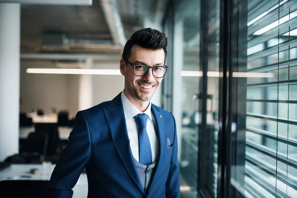 Un ritratto di un giovane uomo d'affari in piedi in un ufficio, guardando la macchina fotografica . — Foto Stock