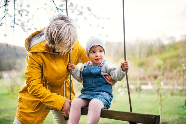 Senior grandmother with toddler granddaughter on a swing in garden in spring. — Stock Photo, Image