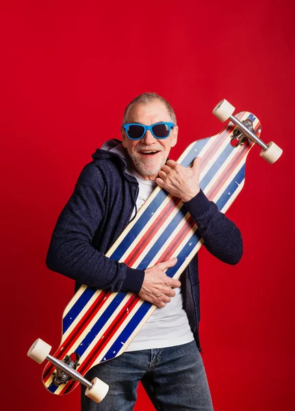 Ein älterer Mann mit Longboard in einem Studio auf rotem Hintergrund, der in die Kamera blickt. — Stockfoto