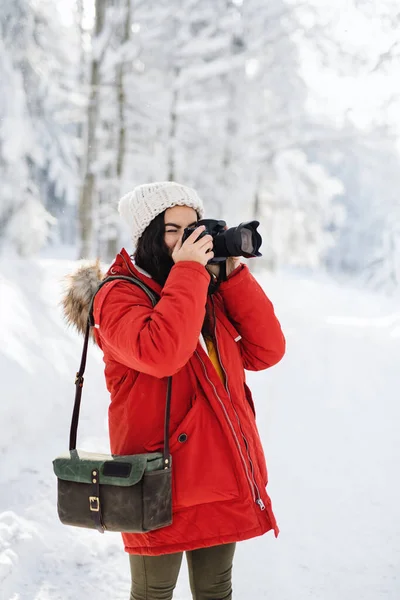Jovem com câmera em pé ao ar livre na neve na floresta de inverno, tirando fotos . — Fotografia de Stock