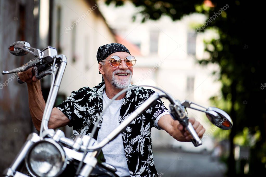 A front view of cheerful senior man traveller with motorbike in town.