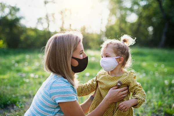 Vista lateral de la madre feliz hablando con la pequeña hija en la naturaleza, con máscaras faciales . — Foto de Stock